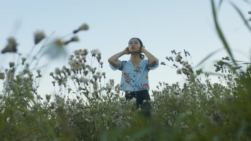 An asian woman stands in a field of wildflowers during the day and puts her hands over her ears
