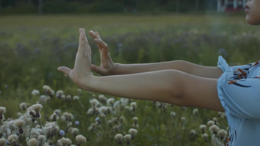 A woman standing and stretching on a grassy field with flowers with a building trees the sky and clouds in the background