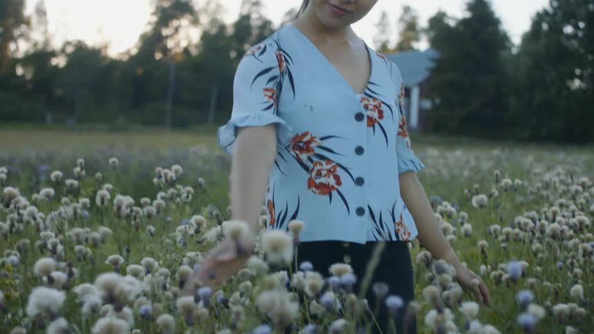 A woman standing on a grassy field with flowers with a building trees the sky and clouds in the background