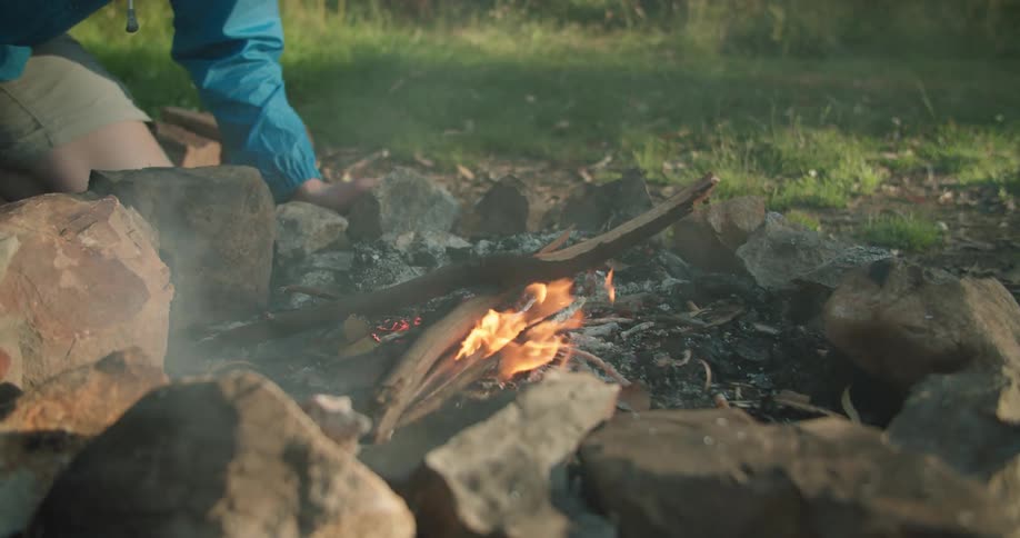 A man kneels to the ground and leans above a burning campfire