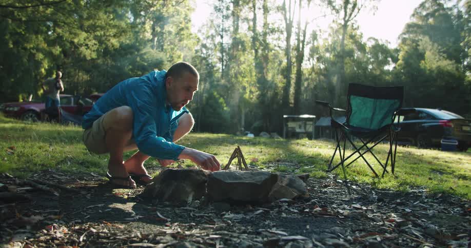 A caucasian man crouched over a campfire trying to get it started in the woods