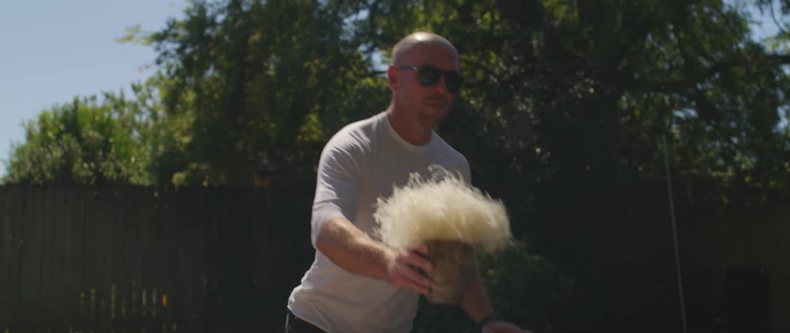 A man with sunglasses placing a plant on a table in a backyard with a fence the sky and clouds in the background