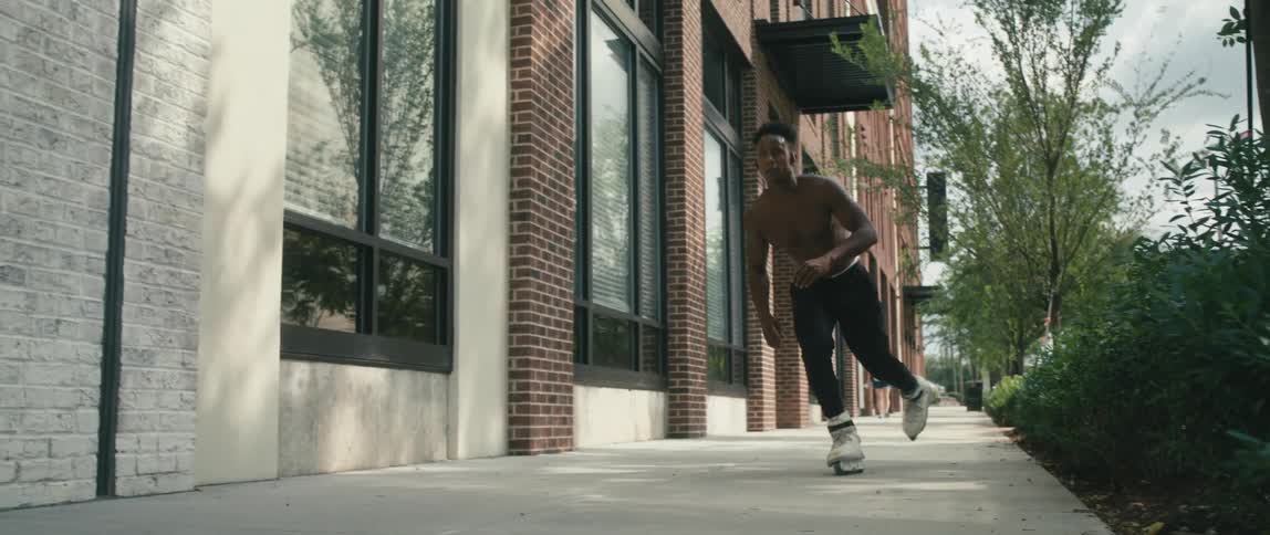 A shirtless man roller blades down a sidewalk past a brick building on a sunny day