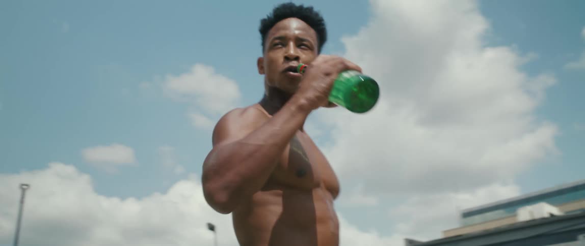 Man sweating with his shirt off takes a sip of a drink while outside on a court
