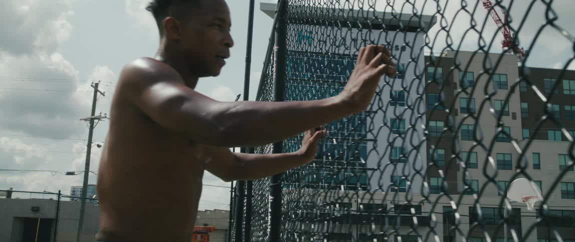 A shirtless black man putting his hands on a metal fence with a hotel in the distance