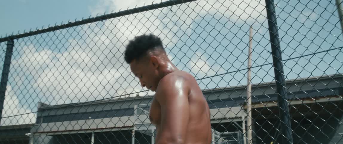 Young muscular fit shirtless man dries sweat from his face with a towel near a wired fence