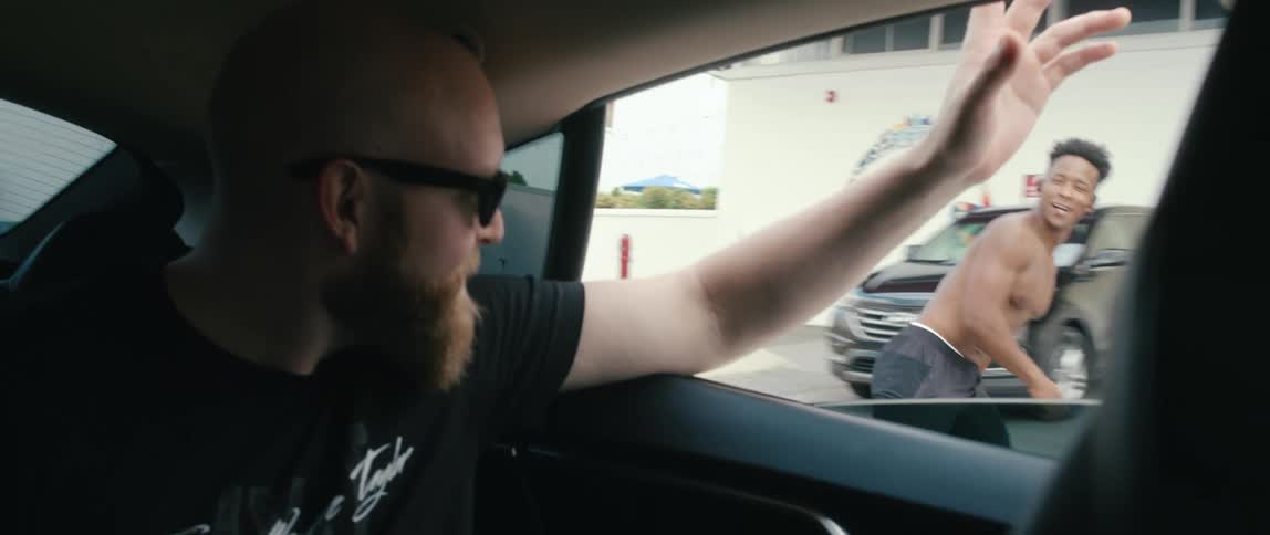 A man riding in a car back seat waves out the window as he passes a roller skater