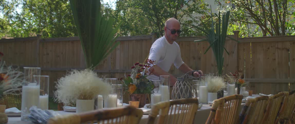 A man arranges tall leaves in a vase on a table in a backyard set for a party