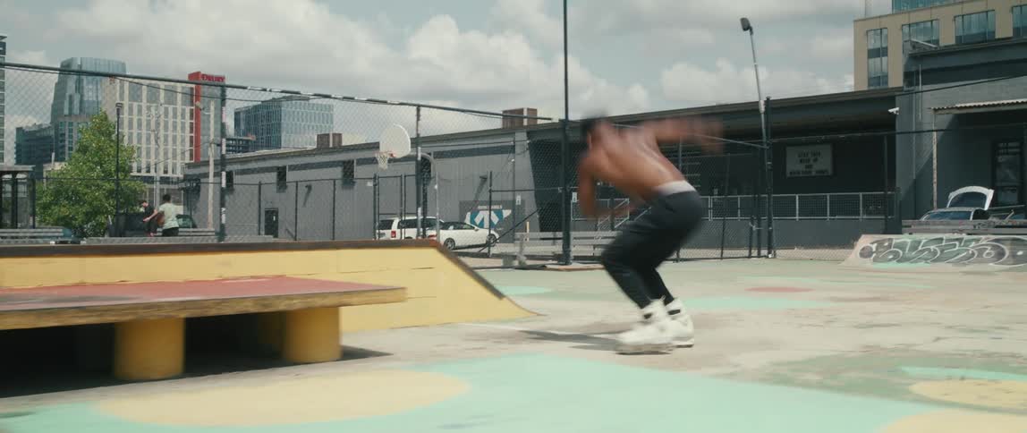 A shirtless teenager roller skates in a skate park nestled between apartment buildings in a city
