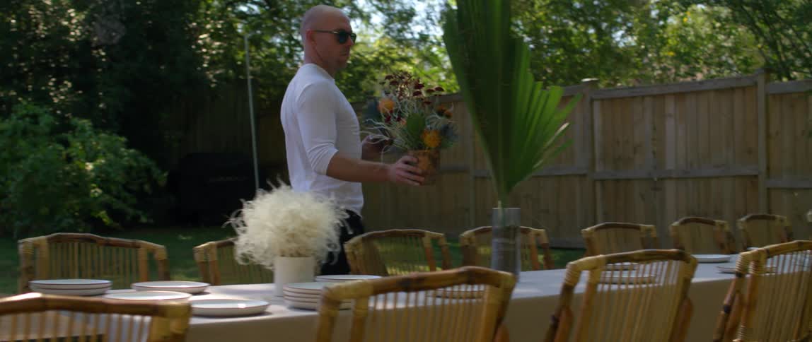 A bald man in a white shirt decorating a table with flower arrangements in the backyard