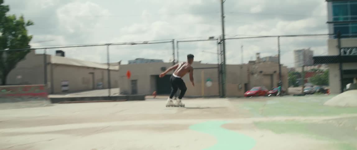 A shirtless young man performs roller blade stunts on the obstacles of an outdoor skatepark in a city during the day