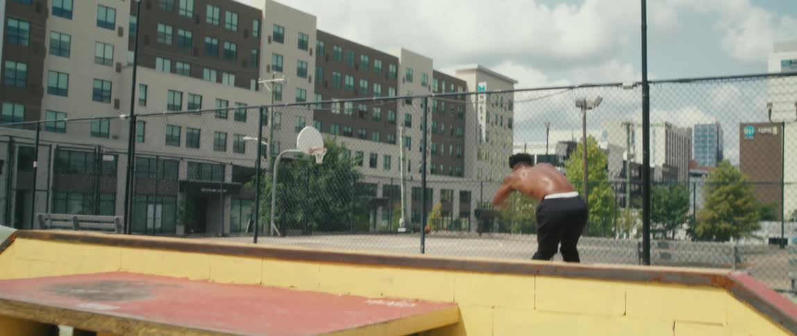 Skater skates up ramp then jumps near basketball court bordered by apartment buildings under a cloudy sky
