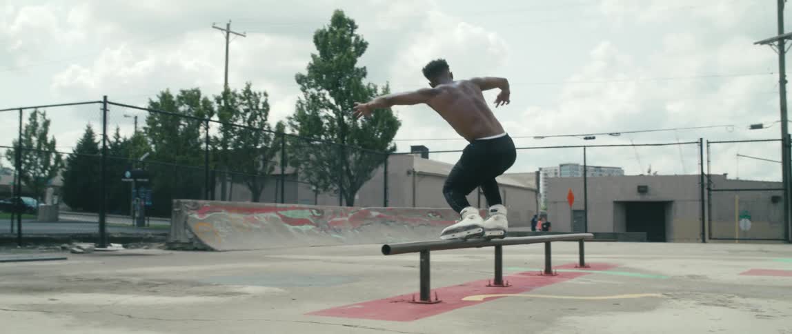 A shirtless young man skating through an outdoor skate park with inline roller skates slides across the top of a rail before jumping off and spinning in the air and landing safely on the ground