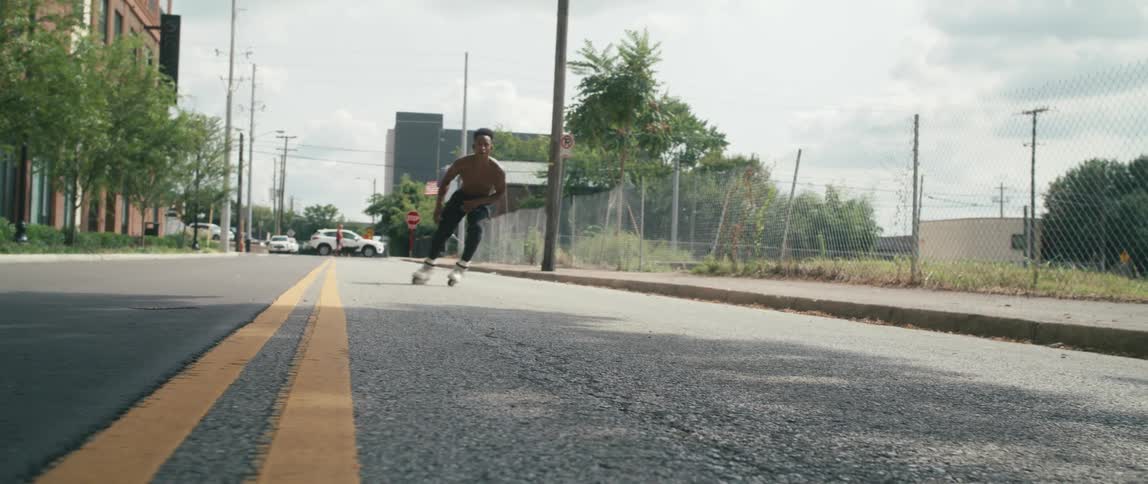 A shirtless young man roller skating down a city road jumps and spins in the air before landing and continuing onward