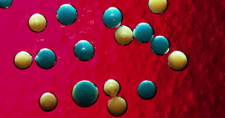 Several droplets of colored liquid are suspended and floating in a clear liquid in front of a red textured surface