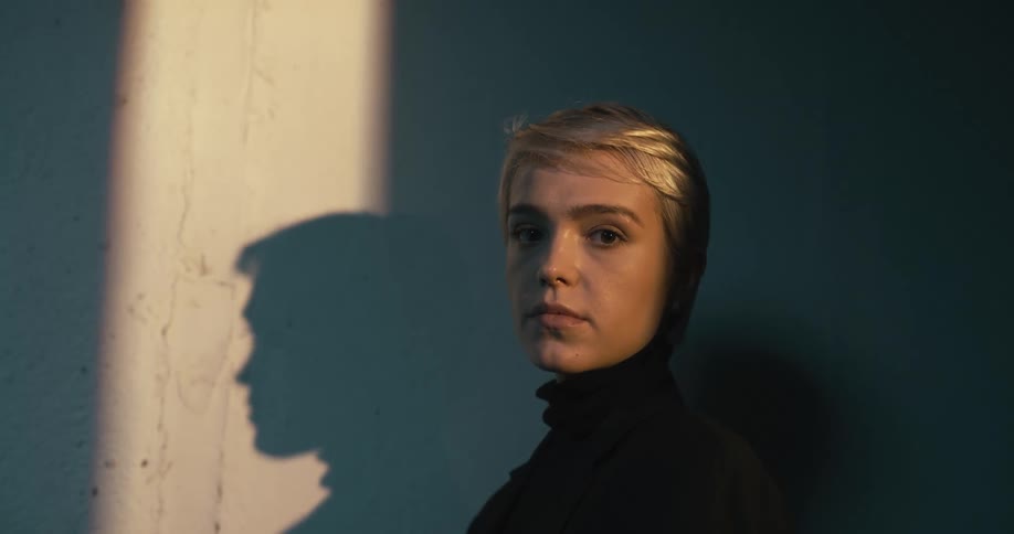 A woman with a turtleneck standing and staring next to a wall with the sun shining on her face