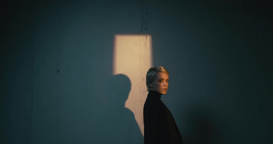 Woman facing to the right inside of a room with a small beam of light turns to look at the camera and then looks away again