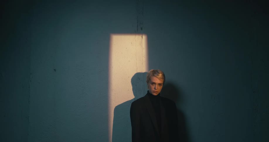 A blonde woman stands against a wall with a block of light on a section