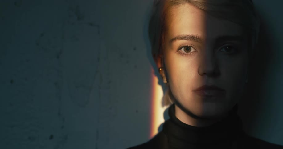 A woman stands in the shadows against a wall with light shining on half her face