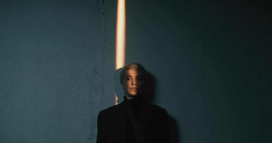 Woman standing with her back up against the wall staring at the camera with a beam of light shining on half of her face