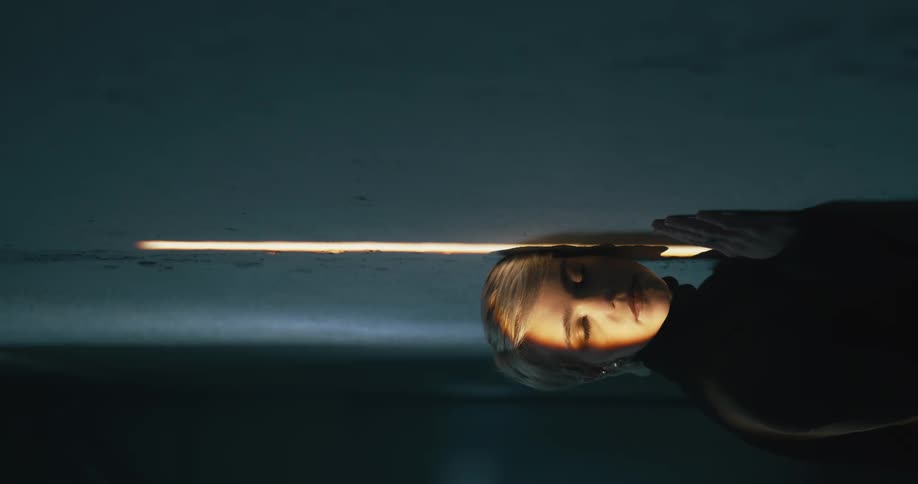 A woman with her eyes closed presses her face up against a wall on a strip of light then opens her eyes to look at the camera