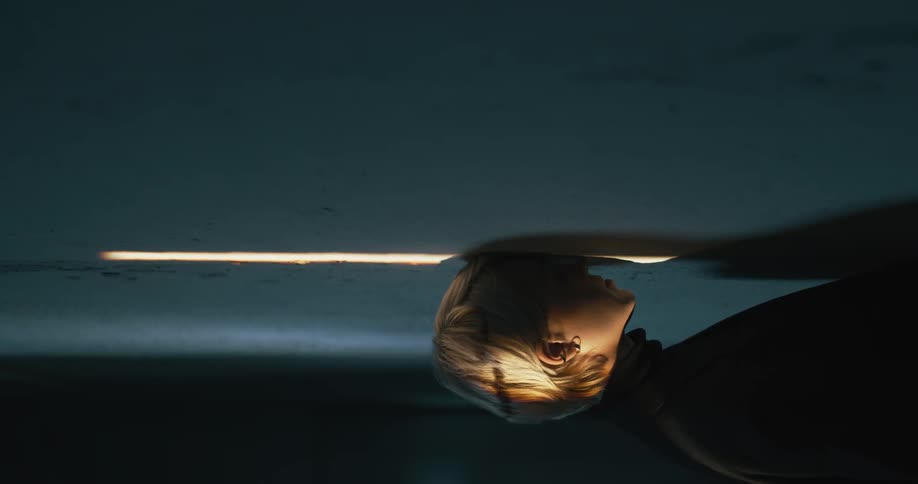 Short haired young blonde woman inside a dimly lit room turns around to look at the camera