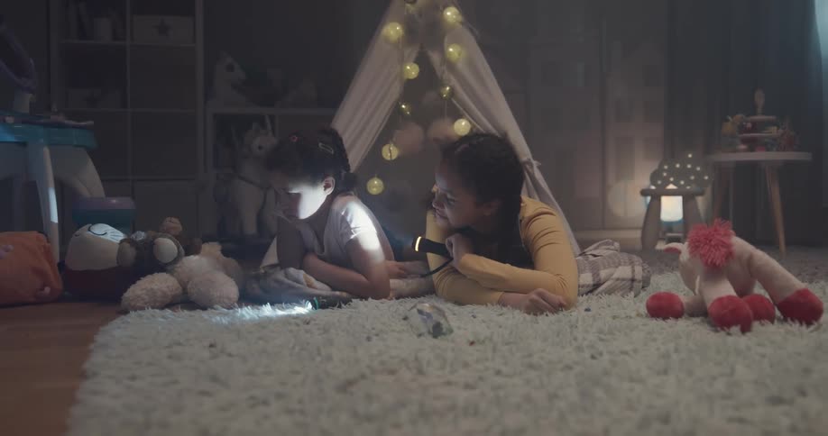 Two young girls pretend to camp out in a room while one shines a flashlight in the other ones face