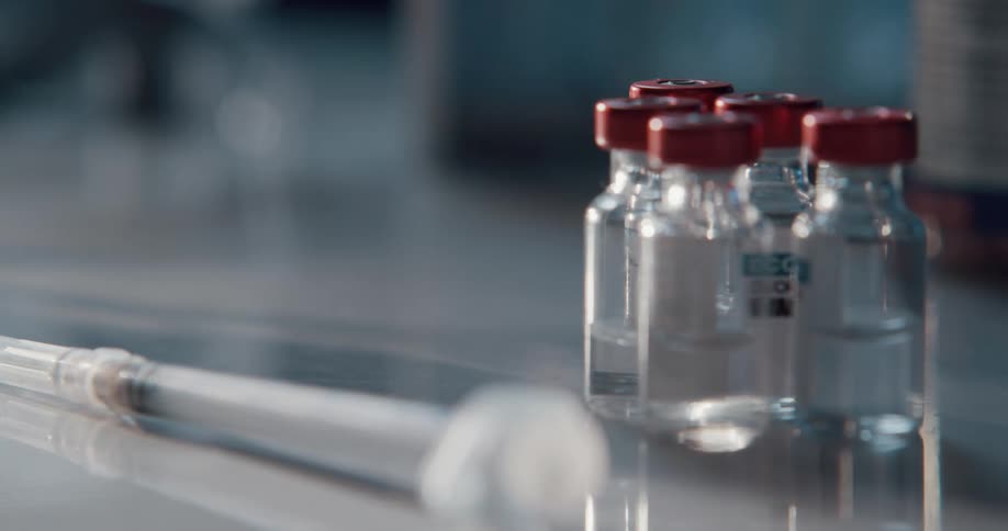 A hand places a needle and a vile of clear medicine on a table next to other vials of medicine