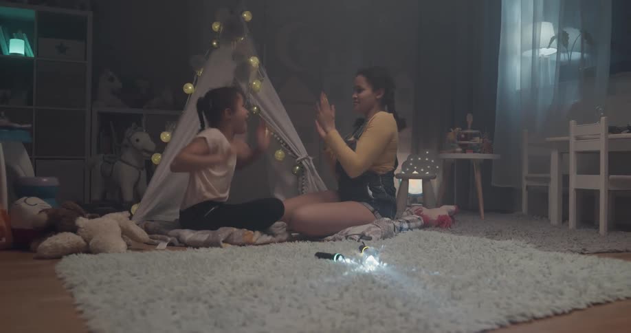 A pair of girls play a clapping game while sitting on a carpet in front of a play tent in a house