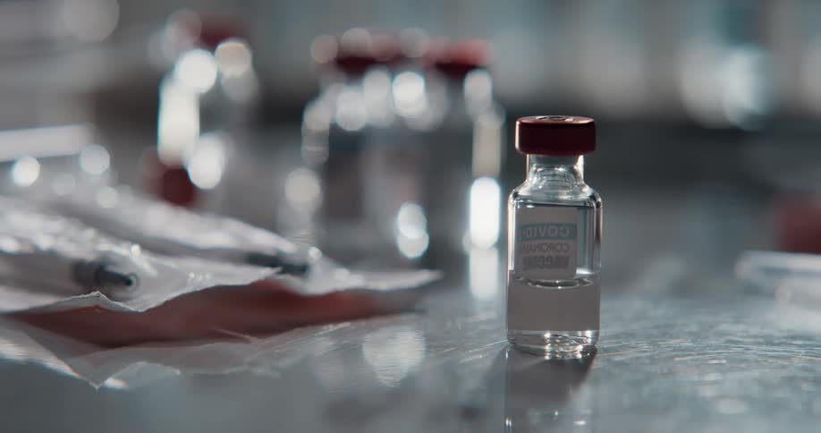 A hand places a vial of a covid vaccine onto a table next to a pack of needles then picks it up again