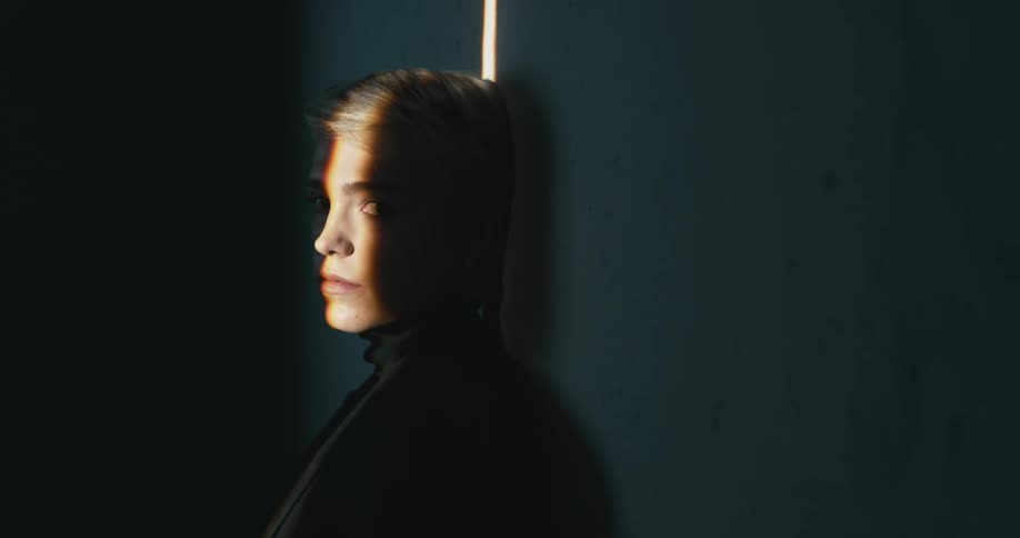 A woman leaning on a wall in the a dark room with a beam of light shining on half of her face looks at the camera