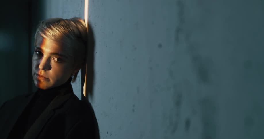 Young blonde short haired woman reclined on a wall modeling for the camera in a dimly lit room