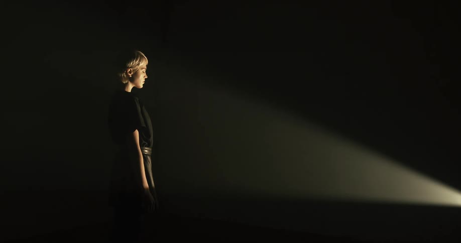 A woman stands motionlessly with her eyes closed in a dark room as a spotlight shines onto the front of her body