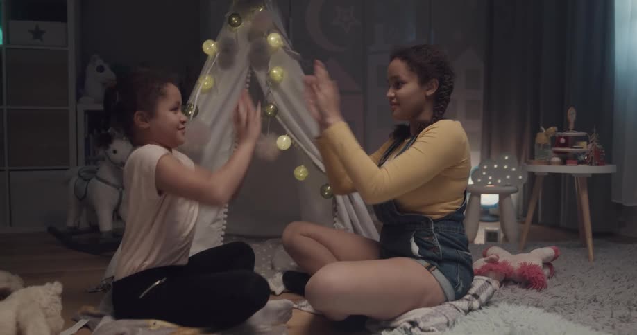 A pair of girls play a clapping game while sitting on cushions on the floor in a house