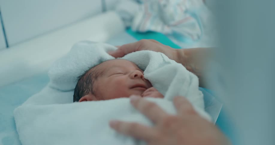 A newborn baby is swaddled in a white blanket being touched by two hands