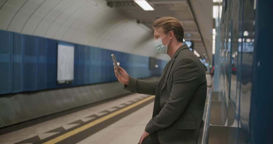 A man using a smartphone while resting in a corridor glances down the hallway before returning his gaze to his phone