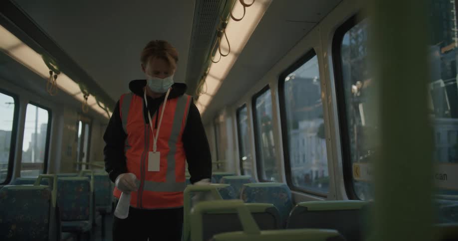 A transit worker wearing a face mask sanitizing seats and railing on a moving train in a city