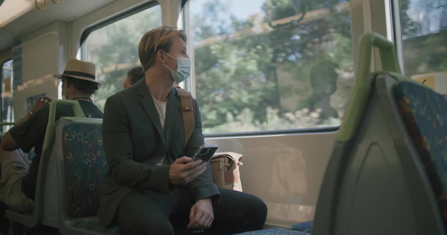 A man with a smart phone and a mask sitting on a train