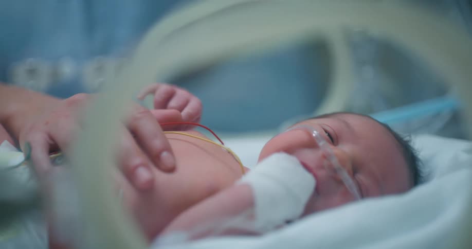 A newborn baby on oxygen lies in an incubator while a woman puts a comforting hand on it