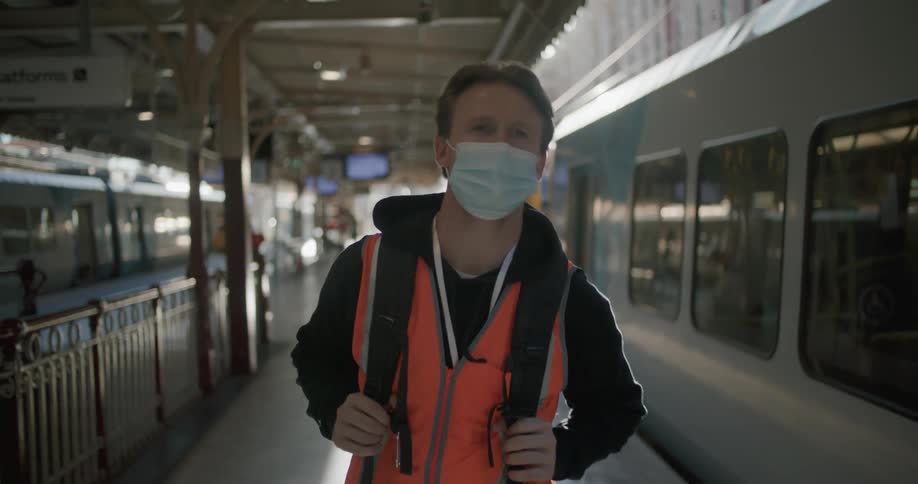 A man with a mask and a backpack walking in a train station with trains