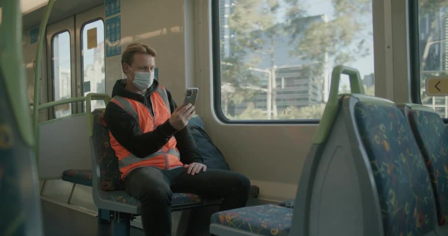 White working man wearing mask and a vest video calling someone on his smartphone while sitting at a train