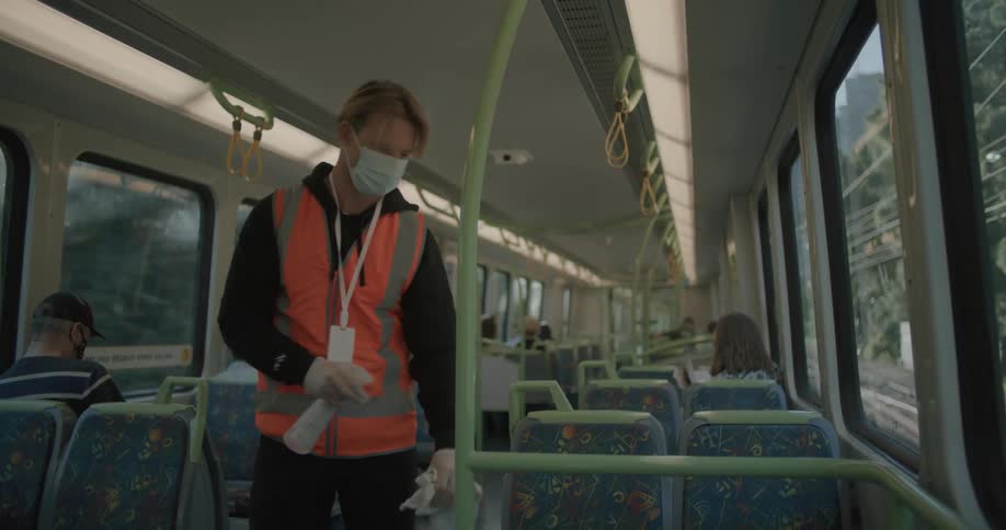 Transit worker wearing a face mask cleaning off hand rails and poles on a moving train at daytime