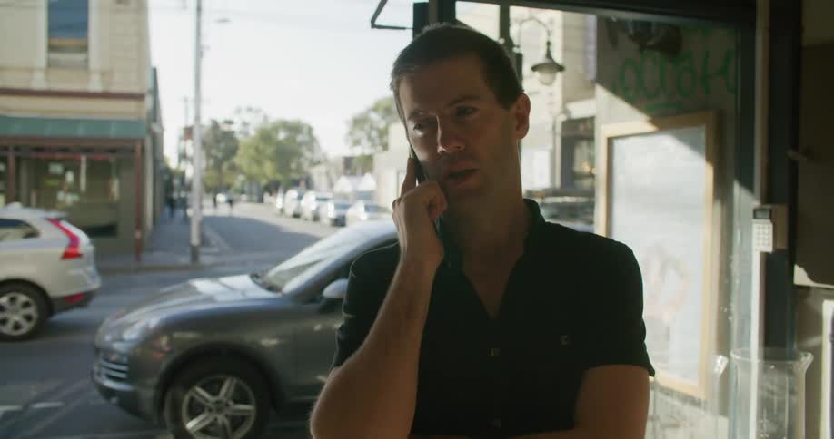 A man talks on a cell phone inside of a building in a city where cars are driving by at daytime