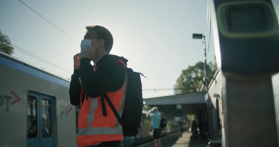 A man wearing an orange shirt and backpack waits on a train platform and places a face mask on his face