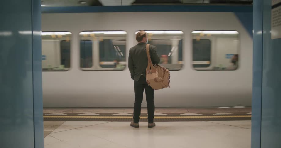 A man dressed in a jacket and pants carrying a tan bag waits on a subway platform