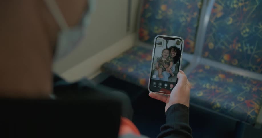 A man waves at his wife and son as he sits on a train and talks to them via video call on his smartphone