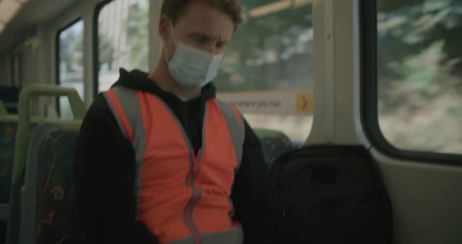 Man wearing safety vest and face mask sits on public transport using mobile phone as transport passes under bridge