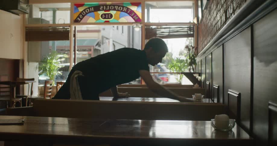 A man sprays and wipes down a table in an empty restaurant as people and cars pass by the open doors