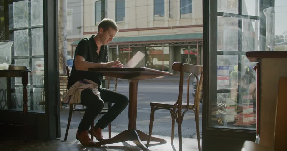 A man sits at a cafe table typing on his laptop next to a busy street with traffic