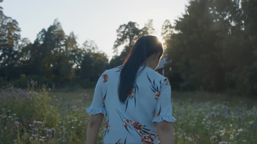 Woman walks through field of wildflowers toward sun shining through trees